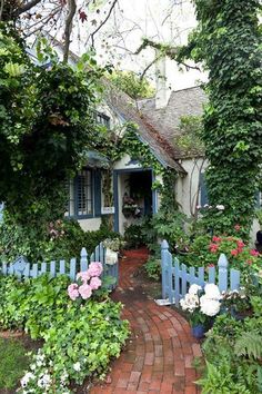 a blue house surrounded by lush green trees and flowers on a brick path that leads to the front door