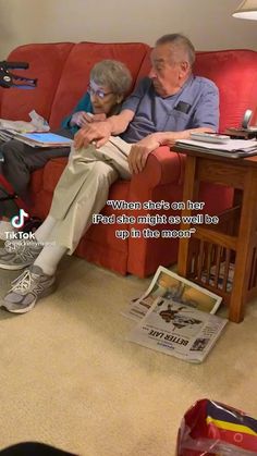 an older man and woman sitting on a red couch in front of a book shelf