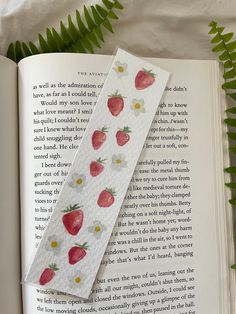 a bookmark with strawberries on it sitting next to an open book and fern