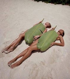 two women laying in the sand on their stomachs, one holding a banana leaf