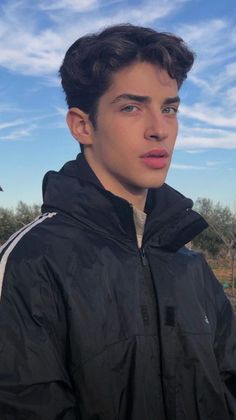 a young man in a black jacket looking at the camera with a blue sky behind him