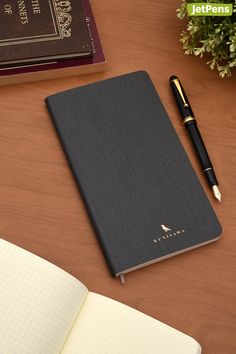 a notebook and pen sitting on top of a wooden table next to a book with writing utensils