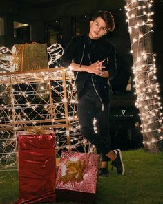 a young man standing next to a pile of presents in front of some christmas lights