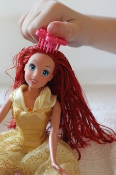 a doll with long red hair is being combed by someone's hand while sitting on a bed