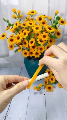 two hands are holding an object near a vase with yellow flowers in it on a white wooden table