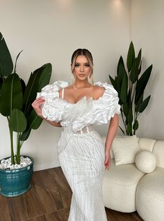 a woman in a white dress standing next to a potted plant and wearing an off the shoulder ruffle gown