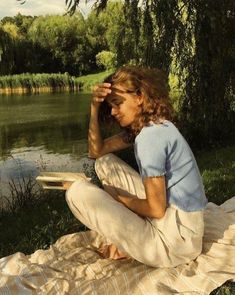 a woman sitting on a blanket next to a body of water with trees in the background