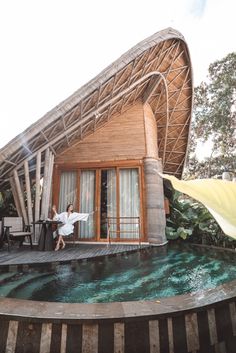 a woman is standing on the edge of a pool in front of a house with a thatched roof