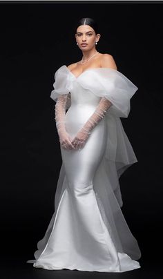 a woman in a white wedding gown with sheer sleeves and gloves on her head, standing against a black background