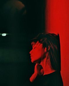 a woman standing in front of a red wall with her hand under her chin and looking off to the side