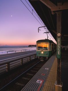 a green train traveling down tracks next to the ocean at sunset or dawn with a full moon in the sky