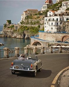 an old car driving down the road next to some buildings and water in front of it