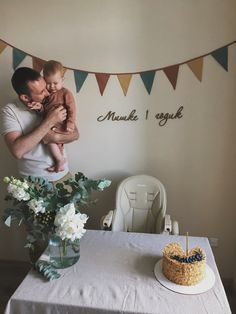 a man holding a baby while standing next to a table with a cake on it