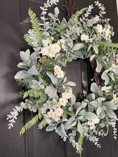 a wreath with white flowers and greenery hangs on a black front door, surrounded by green leaves