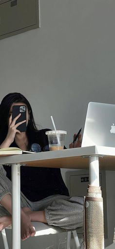 a woman sitting at a table with her cell phone up to her face while looking at the computer screen