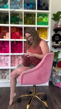 a woman sitting in a pink chair next to a shelf filled with yarn