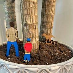 two wooden figures are standing next to a tree in a potted planter with dirt on the ground