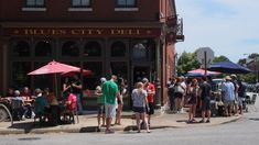 people are standing in front of the blue city deli on a street corner with red umbrellas