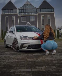 a woman kneeling down next to a white car on a brick road in front of a house