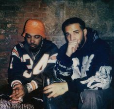 two young men sitting next to each other in front of a brick wall, one with his hand on his face