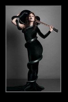 Taylor Swift wearing a long one-sleeved black body con dress holding a black guitar over her head with a long black snake wrapped around her body