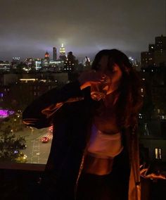 a woman standing on top of a building next to a tall building at night with the city lights in the background