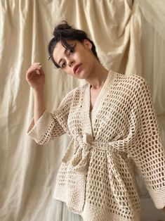 a woman standing in front of a white backdrop wearing a crocheted sweater and jeans