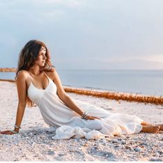 a woman is sitting on the beach wearing a white dress and posing for a photo