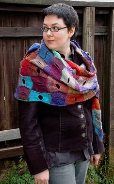 a woman standing in front of a wooden fence wearing a colorful knitted shawl