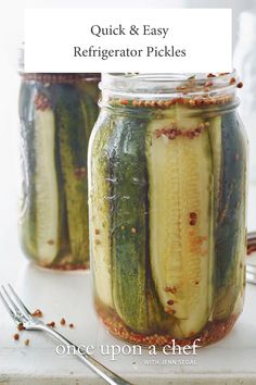 two jars filled with pickles sitting on top of a white counter next to silverware