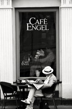 a man sitting at a table in front of a cafe reading a newspaper while wearing a hat