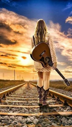 a woman holding a guitar standing on train tracks at sunset with the sun setting behind her
