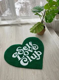 a green heart shaped rug with the words get over club on it next to a potted plant