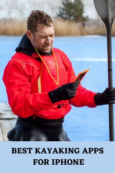 a man in red jacket standing next to a lake holding a cell phone and looking at it