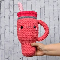 a hand holding a pink crocheted coffee cup with a face drawn on it