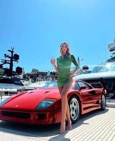 a woman standing next to a red sports car on a boat dock with other boats in the background