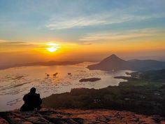 a person sitting on top of a mountain watching the sun go down over a body of water