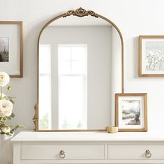 a white dresser topped with a mirror next to a vase filled with flowers and framed pictures