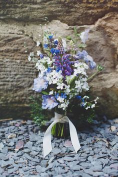 a bouquet of blue and white flowers sitting on top of a gravel ground next to a stone wall