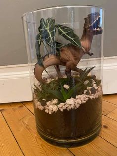 a glass vase filled with fake dinosaurs and plants on top of a wooden floor next to a wall