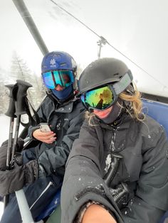 two people sitting on a ski lift in the snow with goggles on their heads