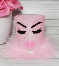 a pink cup with black eyes and eyelashes on it next to a white vase filled with pink flowers