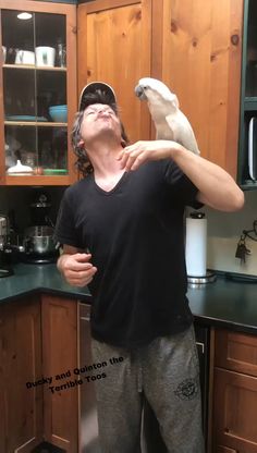 a man standing in a kitchen with a dog on his shoulder and looking up at the sky