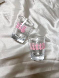 two shot glasses sitting on top of a white bed covered in pink fabric with the words, big sister and little sister printed on them