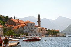 people are sitting on the dock near boats and buildings in the water with mountains in the background