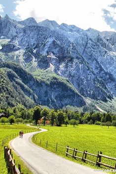 two people riding horses down a country road in front of some mountains and green grass