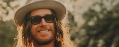 a man with long hair wearing sunglasses and a hat smiles at the camera while standing in front of trees