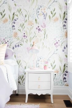 a bedroom with floral wallpaper and a white nightstand in front of the bed, next to a window