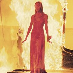 a woman in a red dress standing on a stage with fire behind her and an orange background