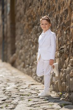 a young boy in a white suit leaning against a stone wall with his hands on his hips